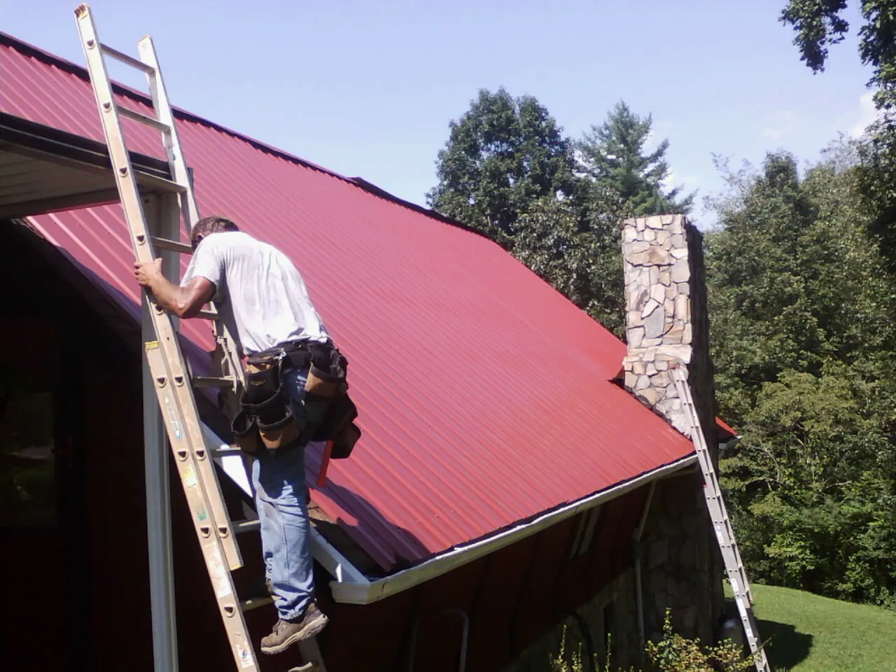 Roofer on a ladder beside a red metal roof and stone chimney — Soffit & Fascia Repair by Roofing Brothers in Tuckasegee